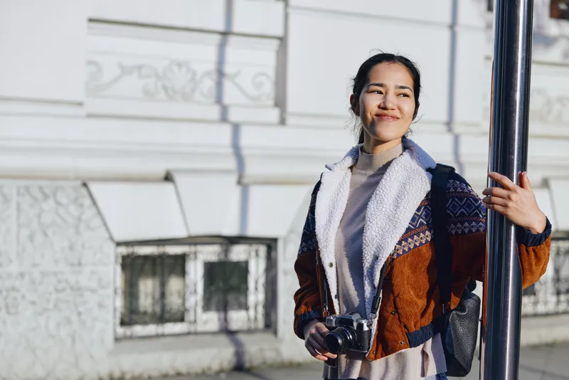 Woman with camera outdoors in urban fashion style, scarf and sweater, jacket against street and pole, bright sunlight, candid moment capturing streetwear vibe and travel mood