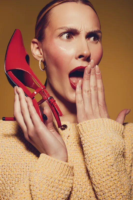 Surprised woman in yellow sweater holds red high heel shoe near face with open mouth and wide eyes. Concept of fashion, shock, style, and unexpected emotions in studio lighting.
