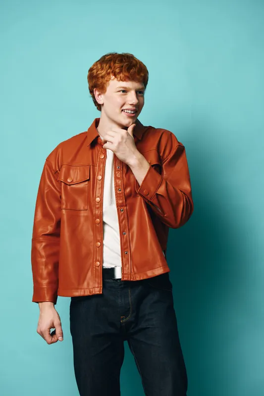 Young man with red hair wearing brown leather jacket and white shirt stands smiling on blue background, casual style with thoughtful pose for fashion or lifestyle portrait.