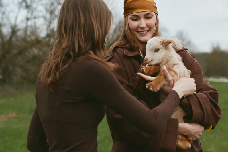 Goat kid with women outdoors, lifestyle, friendship, nature, countryside, warm scene of care and bonding in rustic setting, candid moment that conveys affection, calm mood, peaceful field