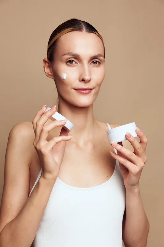 Young woman applies moisturizing cream on face holds jar of skincare product looks at camera with gentle smile isolated on beige background natural beauty concept closeup portrait