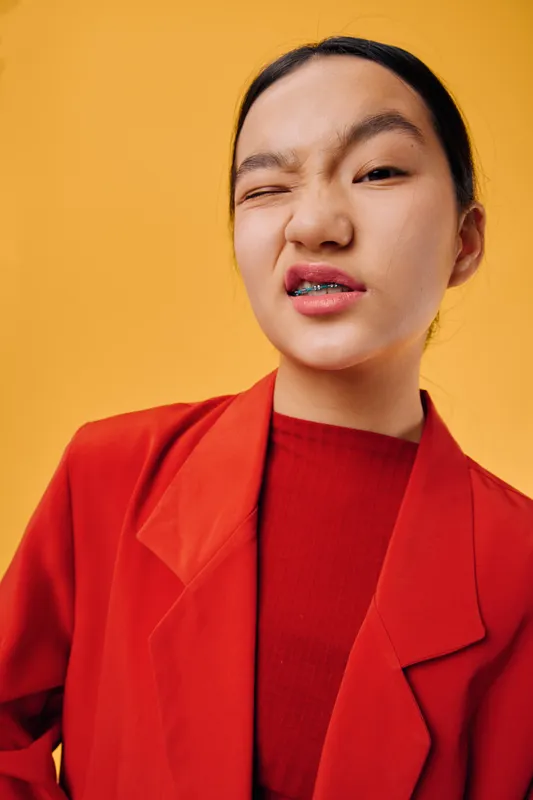 Young woman in red blazer and shirt winking with confident expression on yellow background. Portrait of female model showing attitude and style in casual fashion pose.