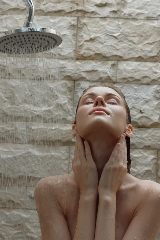 woman showering under rain showerhead with relaxing expression, stone wall background, wet skin, wellness ritual, natural light