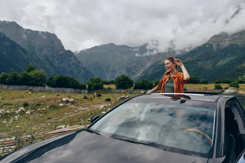 Person leaning out of a sunroof in a car during a mountain landscape road trip, enjoying fresh air and expansive scenery in a rural setting.