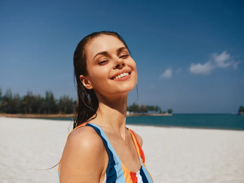 Sunny portrait of a smiling woman on a bright beach, warm sun, clear sky, and calm blue sea, capturing carefree summer mood and natural confidence.