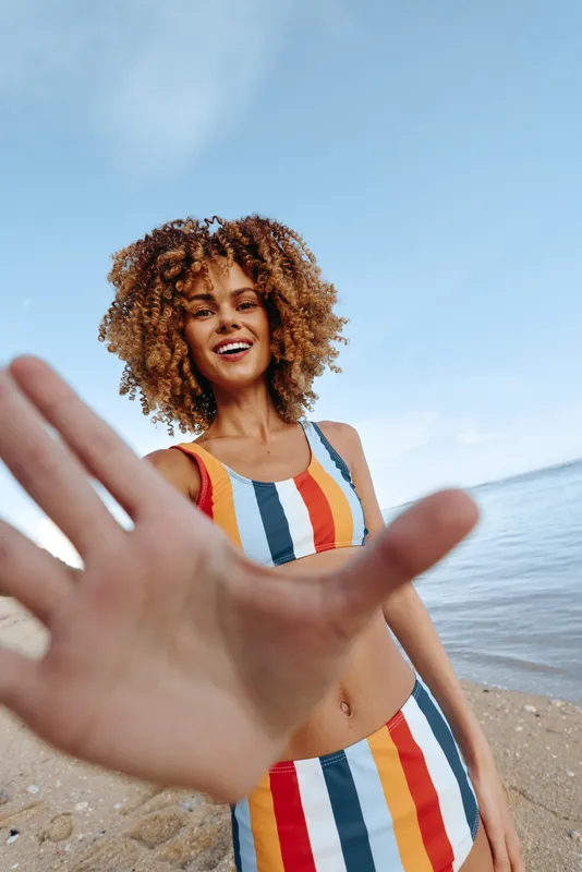 Cheerful woman on a sunny beach, wearing a colorful striped swimsuit, reaching toward the camera with a smile, blue sky and calm ocean in the background, relaxed summer vibe