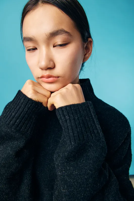 Young woman in black sweater with closed eyes expresses sadness and contemplation on blue background. Portrait, emotion, and fashion with natural light and calm mood.