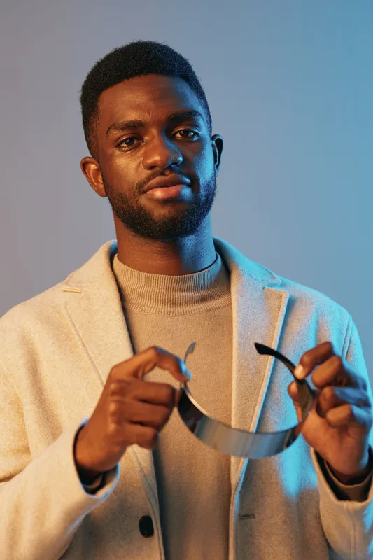 Confident young male model holding a modern accessory, dressed in a stylish beige coat against a cool blue background, embodying urban fashion and confidence