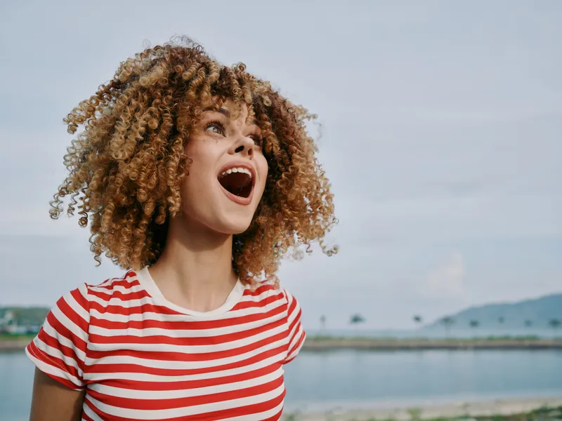 Joyful young woman with curly hair wearing a red and white striped tee stands by a lake, laughing with an open mouth as wind lifts her curls under a bright sky.