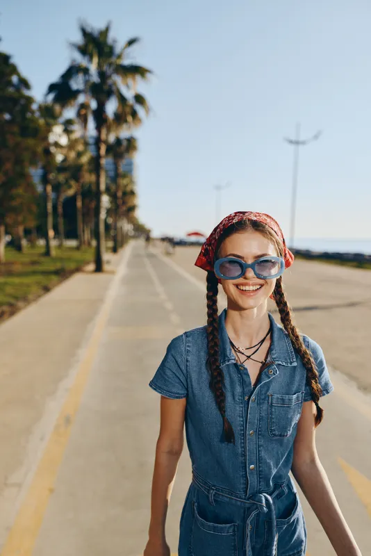 Lifestyle woman in modern boho-western denim street style wearing sunglasses and bandana walking outdoors, smiling in warm natural light with film color effect.
