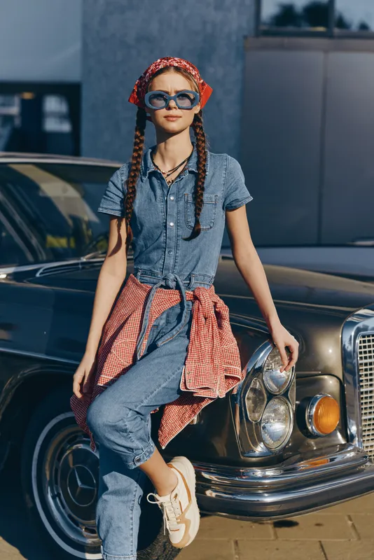 lifestyle woman in modern boho-western denim street style with braided hair and sunglasses leaning on vintage car, film color effect enhancing casual urban vibe
