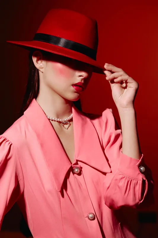 Fashionable woman wearing a red wide-brimmed hat and pink textured jacket closes her eyes with hand touching hat brim. Creative beauty editorial concept with vibrant colors and elegance.
