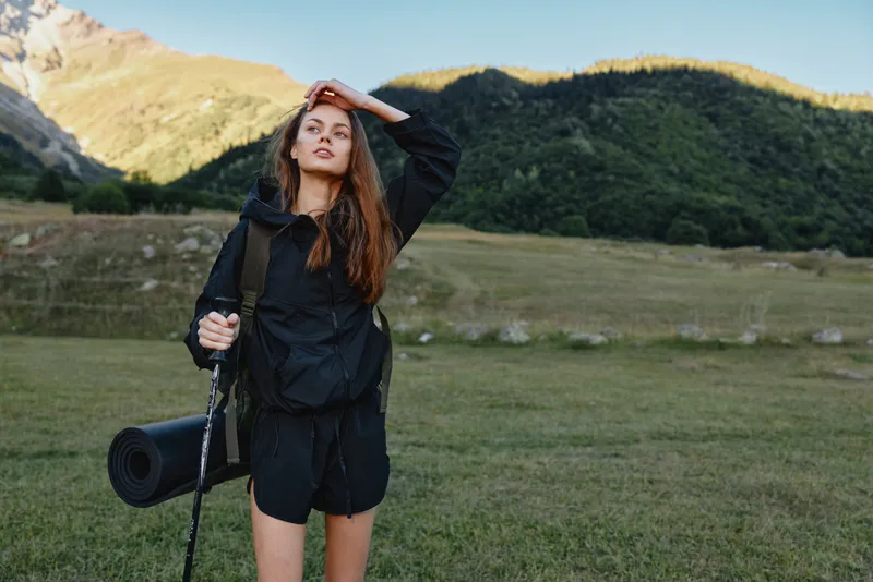 A confident outdoorswoman stands in a wide valley with mountains in the background. She wears a black jacket and shorts, carrying a yoga mat and backpack, ready for adventure.
