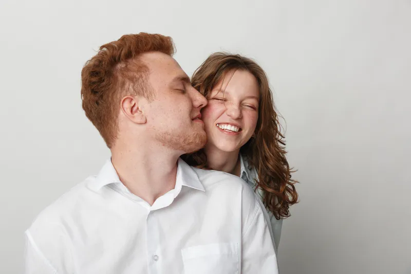 Happy young couple in casual clothes share a tender moment, man kissing woman on cheek. Bright studio background, concept of love, affection, and relationship happiness.