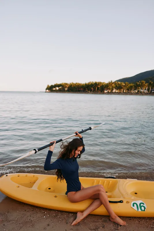 A person sits in a yellow kayak on calm water by a sandy shore, raising a paddle overhead as a playful, athletic moment unfolds against a clear sky.