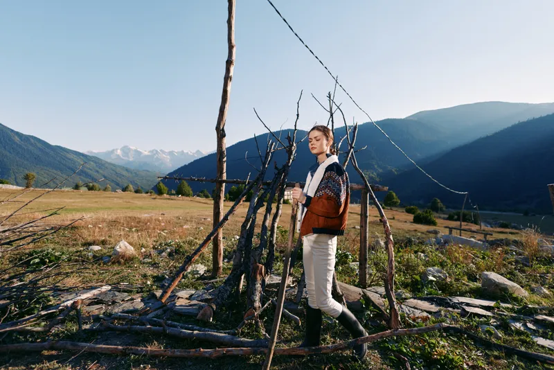 woman mountains field cup sweater rural nature standing by a rustic stick fence in an alpine valley, cozy jacket and boots enjoying morning sunlight and scenic mountain landscape
