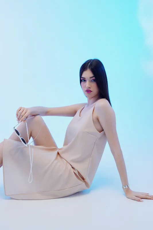 Young woman beauty fashion model with long dark hair wearing a beige dress sitting on the floor against a blue background. Concept of elegance and style.