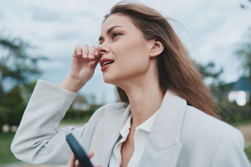Woman in white jacket thoughtful expression outdoors holding phone with cloudy sky background, natural light, autumn season, casual fashion style