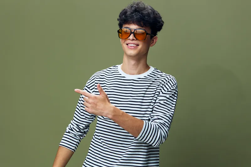 young man wearing striped shirt and sunglasses smiling confidently while pointing sideways on olive green background, casual style, positive mood