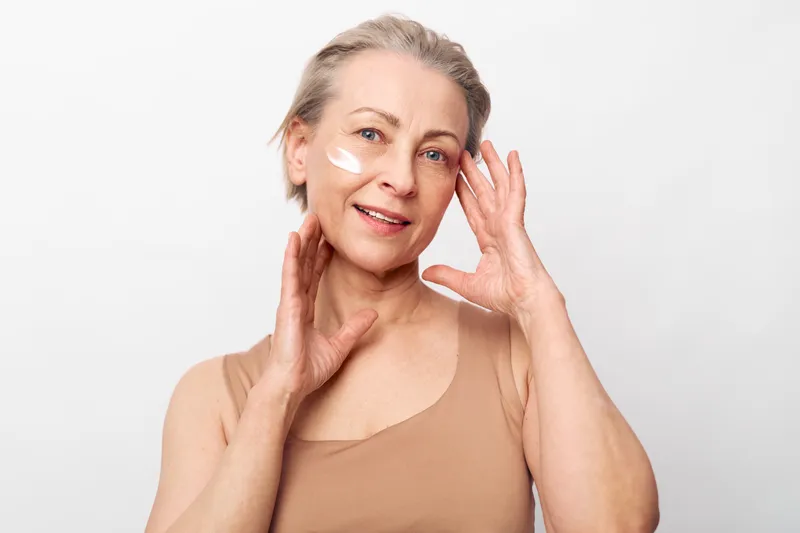 Mature woman with light skin applies white face cream on cheek smiling gently against plain light background. Concept of skincare, aging beauty, self-care, wellness, and natural cosmetics.