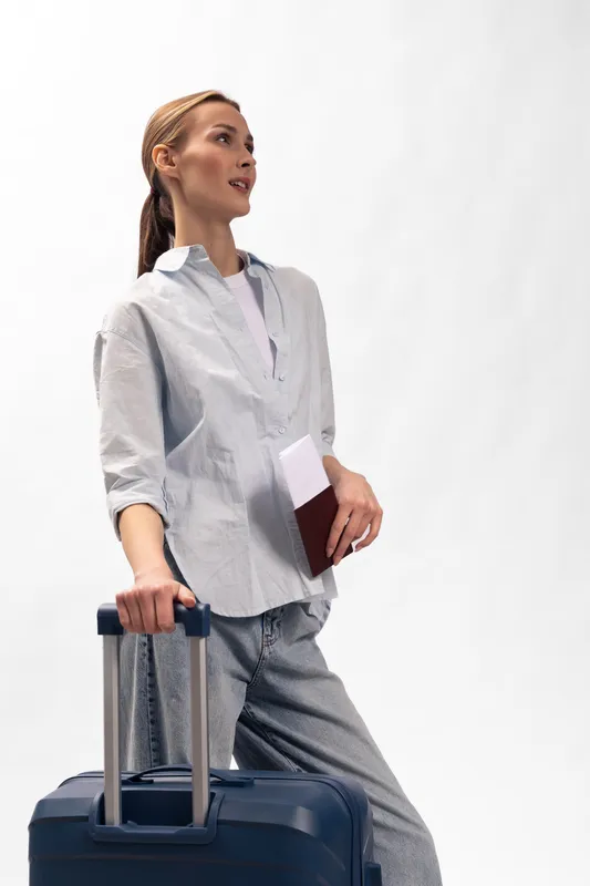 Young woman with ponytail holding travel documents and suitcase handle looks away. Casual outfit and minimal background create travel and vacation concept with relaxed mood.