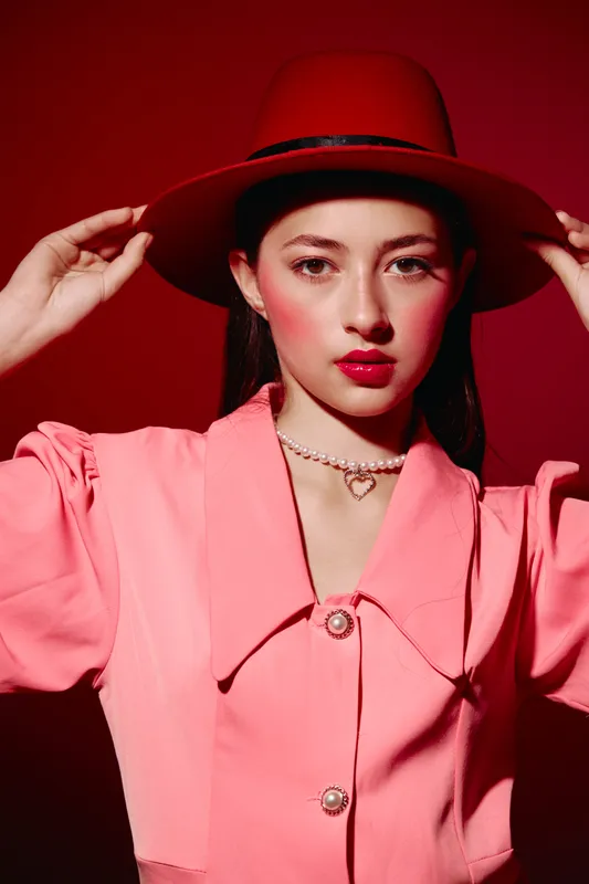 Young woman wearing a red hat and bright pink coat poses against red background with bold makeup and pearl necklace. Creative beauty fashion portrait showcasing style.
