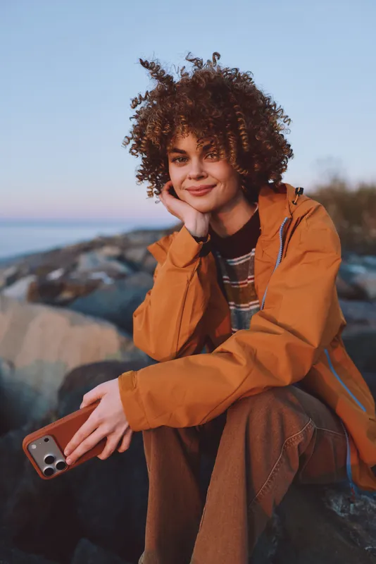 Woman with curly hair smile in orange jacket outdoors, holding phone. Mountains background at sunset, lifestyle travel mood, soft wind, natural portrait, casual fashion in fresh air.