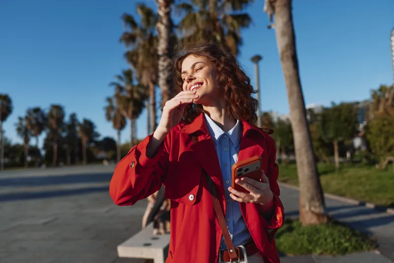 Young woman with curly hair smiles outdoors in a bright red coat and blue shirt, holding a phone and enjoying a sunlit moment on a palm lined street.