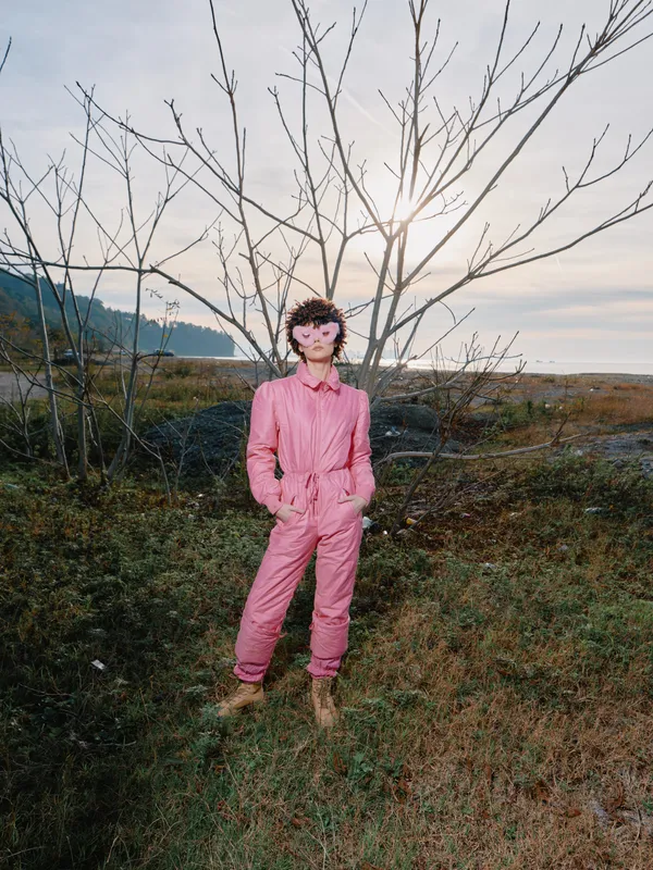 Person wearing pink jumpsuit stands in an open field with bare trees and a wide landscape, casual street style in nature, solo portrait under daylight, muddy ground, distant hills, soft clouds above.