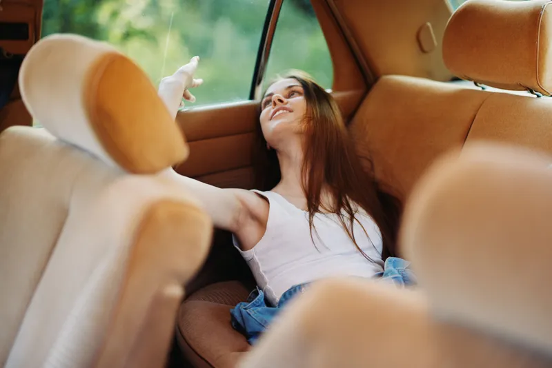 Young woman relaxing in a car, wearing casual clothing, surrounded by soft seats, exuding a carefree and joyful emotion, showcasing a serene outdoor background.
