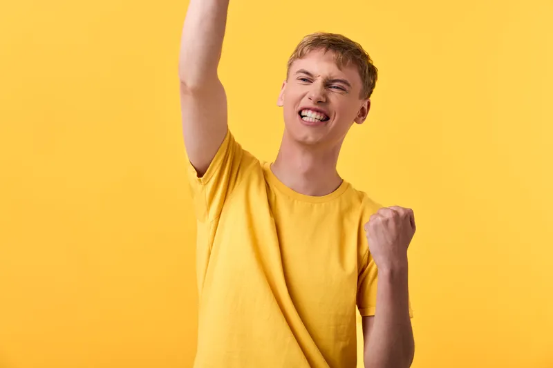 Man in a bright yellow shirt raises a clenched fist in celebration, capturing triumph, excitement, and pure energy against a bold yellow studio background.
