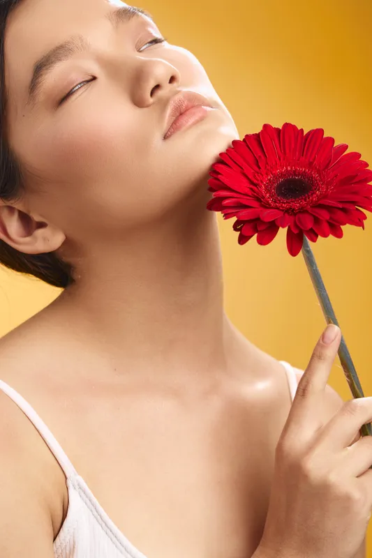 asian woman cradles a red flower near her cheek, eyes closed, serene expression, soft lighting, warm background, graceful portrait conveying beauty and calm.