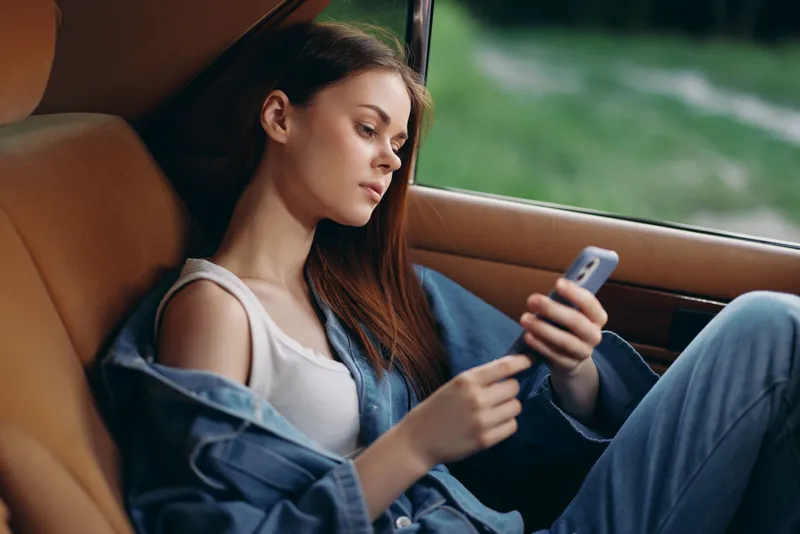 Young woman relaxing in the car, wearing casual clothing, focused on her phone, showcasing a serene expression in a natural green background.