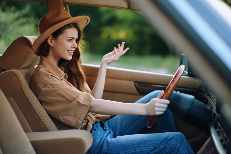 A young woman with long hair, wearing a brown hat and a casual outfit, joyfully driving a vintage car in a lush green setting, depicting a sense of freedom and adventure.