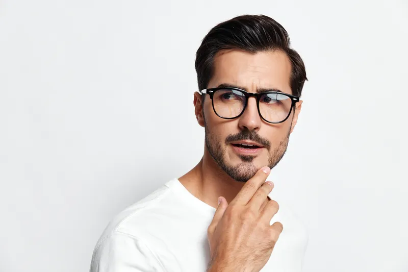 Thoughtful man wearing glasses and a white t-shirt, with a curious expression on his face, looking away while pondering over a question in a bright studio setting.