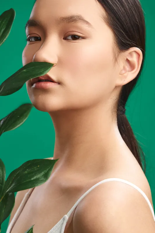 asian woman in a clean studio portrait with lush greenery framing her face. she gazes softly, conveying calm confidence and a fresh, natural mood ideal for contemporary beauty and fashion.