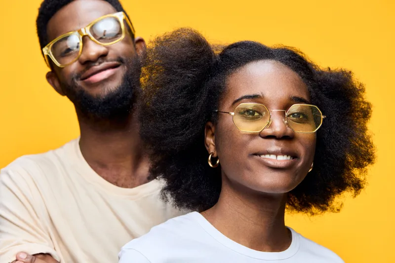 Young diverse couple with stylish glasses against a vibrant yellow background, expressing joy and confidence in contemporary fashion and urban style