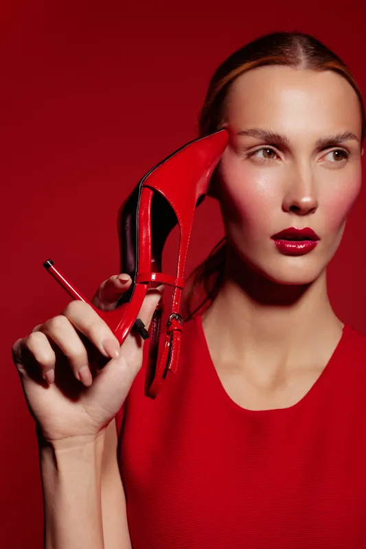 Stylish woman in red dress holds red high heel shoe near face on red background. Fashion concept with bold makeup and elegant pose for beauty and style promotion.