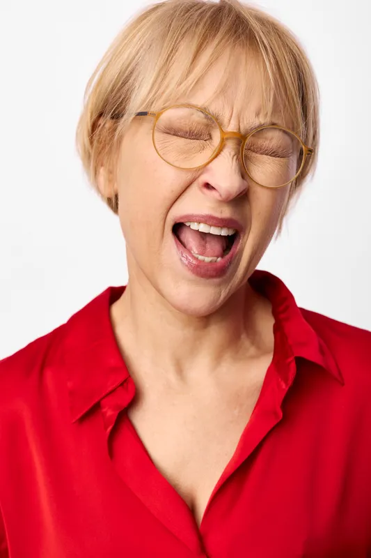Cheerful blonde woman, laughing with eyes closed behind round glasses, open mouth smile, red shirt, studio portrait on white background, joyful mood