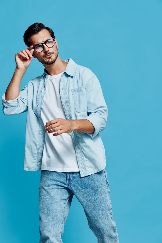 Stylish young man wearing light blue shirt and glasses poses confidently against a vibrant blue background, showcasing a modern and fashionable look.