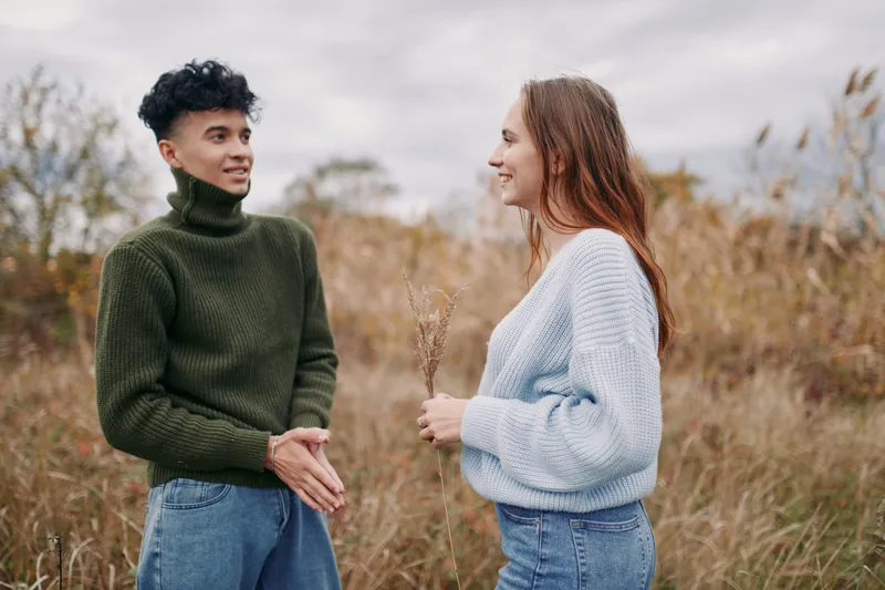 Young couple sharing a candid outdoor conversation in a natural field, conveying authenticity and trustworthiness through genuine connection, relaxed smiles, casual sweaters and warm, honest