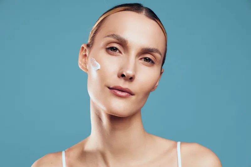 Young woman with clear skin applies white cream on her cheek while looking calmly at the camera against a plain blue background.
