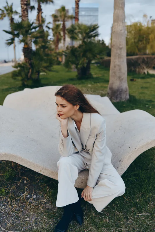 Business woman in a stylish suit, seated on a modern bench outdoors, contemplating her next move amidst palm trees and greenery.
