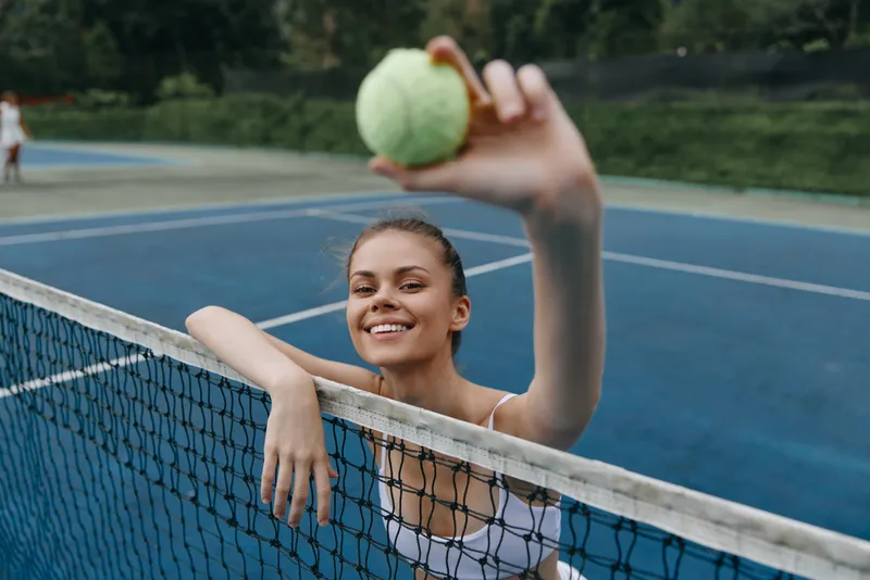 Young woman playing tennis, smiling and holding a tennis ball at the net, showcasing enthusiasm and sportsmanship on a blue court Tennis, sport, smile, blue, young woman, ball, net
