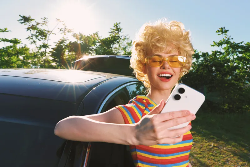 Cheerful woman with curly blonde hair in a bright striped top takes a selfie by a car, sunny outdoor scene with retro film color and casual lifestyle mood and warm glow adds nostalgia.