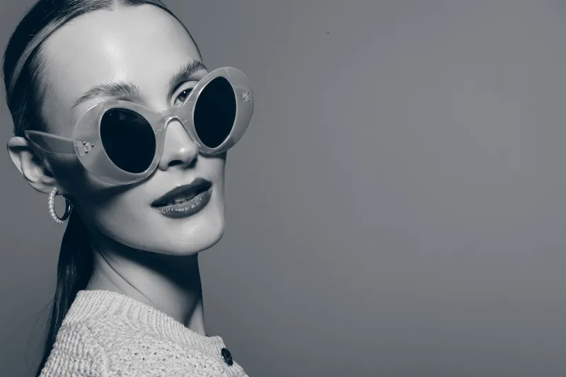 Stylish white woman with light skin wearing round sunglasses and hoop earrings looks confidently in studio. Concept for fashion, glamour, and portrait photography.