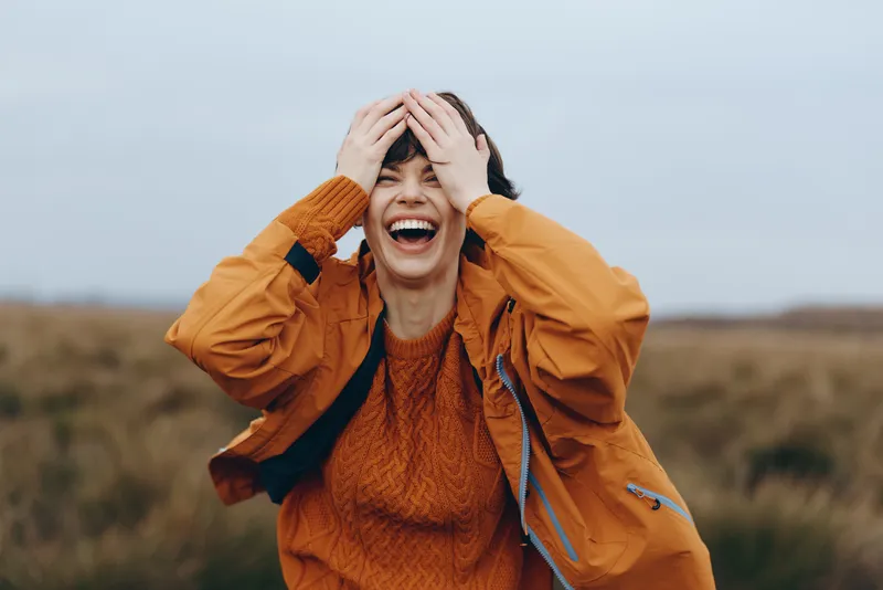 Woman laughing outdoors in an orange coat, hands covering her eyes in a countryside field. Windy overcast sky creates a lively lifestyle moment, natural daylight, candid expression and joy.