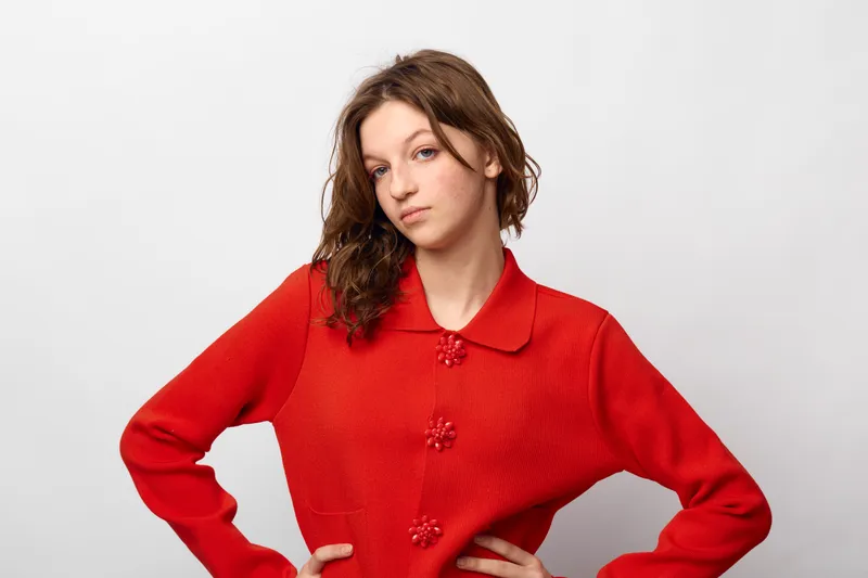 Young woman with wet hair wearing a red sweater stands with hands on hips against white background. Casual style, confident pose, and natural expression in studio lighting.