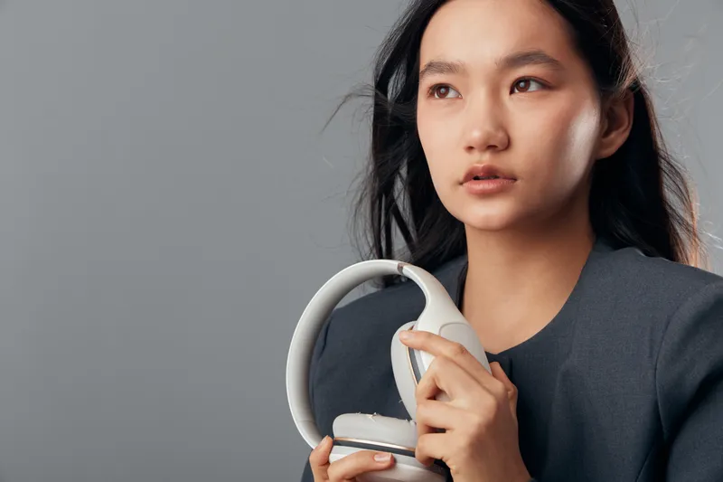 Young woman holding white headphones looking away with serious expression isolated on gray background. Portrait of girl with long dark hair preparing for music or audio experience.