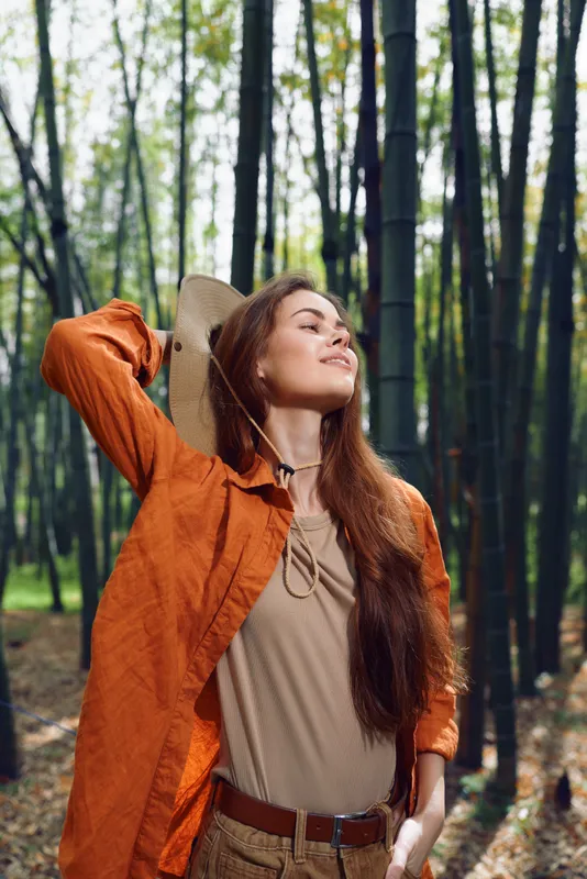 Woman in orange jacket and hat stands in bamboo forest, smiling with eyes closed in a nature portrait, serene autumn sunlight on longhair for outdoor travel and leisure.